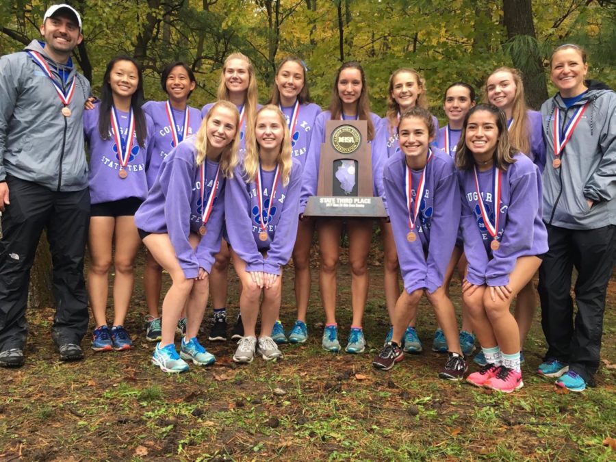 This photo shows the girls cross country team and their coaches posing for a picture with their trophy at state. The girls are all wearing purple sweatshirts and medals.