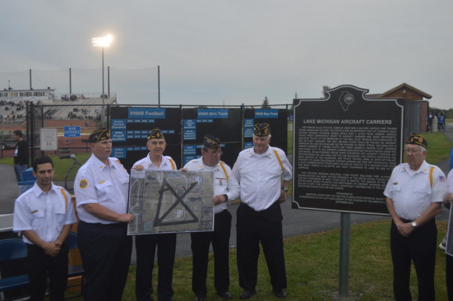 This is a picture of 6 military veterans dressed in all white standing next to a plaque in front of the football field.