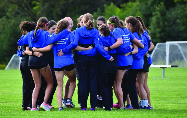 A large number of girls on the varsity cross country team huddle together in a circle with their arms around each other.