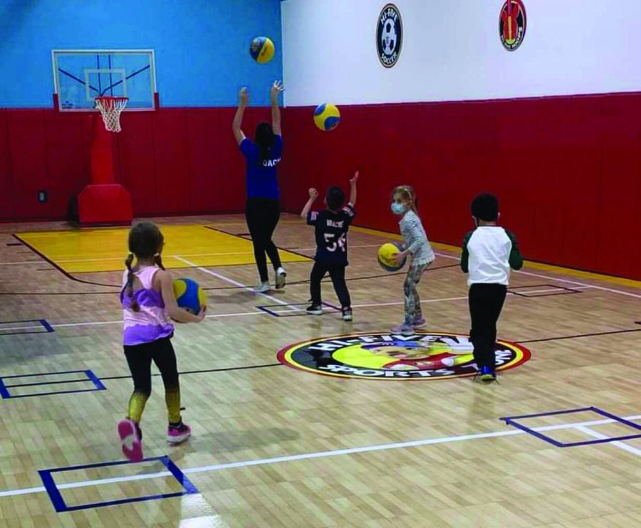 Marianna leaps as she tosses her basketball into the basketball hoop. There is a line of three kids behind her preparing to shoot their basketballs as well. One small girl is running towards the back of the line with her ball.