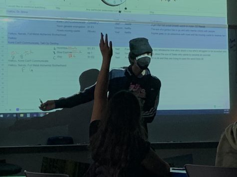 Jonathan Aguila stands in front of a screen projecting an image of a list of animes. Jonathan points to numbers in Japanese characters as he counts votes for what anime to watch during an Anime Club meeting. In front of him, a student raises their hand.