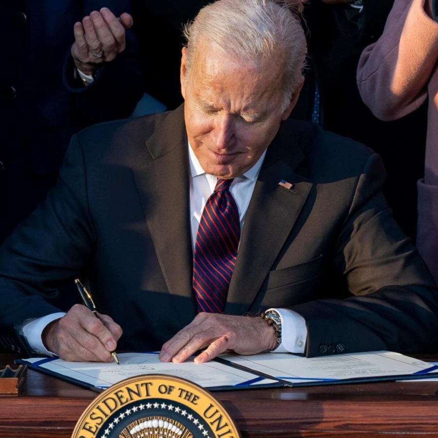 Joe Biden sits at a desk while signing a document. The desk has the presidential seal on it.