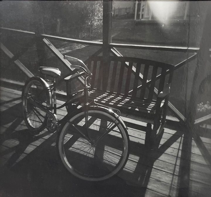 Black and white film photograph of a bike leaning on a bench sitting on a bridge.There are sun beams shining through the bench and bike.